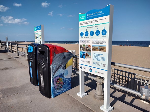 A sidewalk with two bins and two signs in front of a beach.